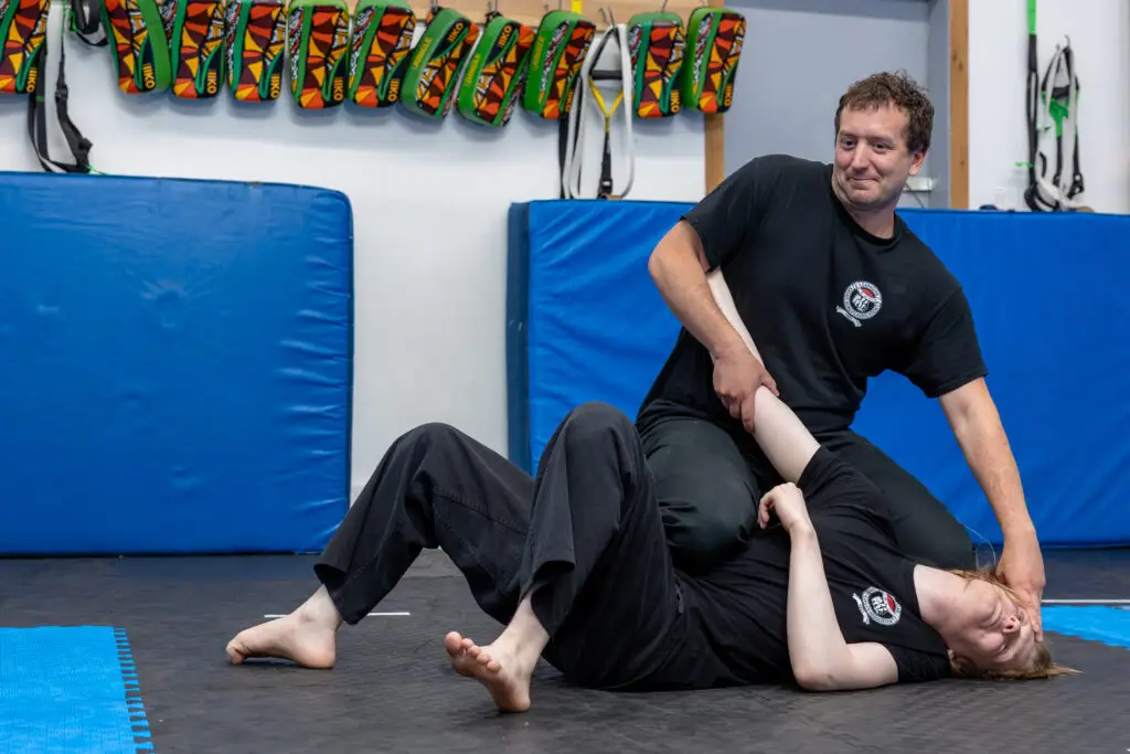 Two people in black shirts grappling on blue gym mats with wall padding and colorful gear hanging in the background.