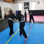 Group of students in black uniforms practicing martial arts on blue mats in a dojo, legs visible and belts tied around waists.