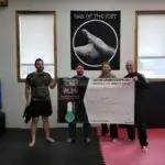 Four people in a gym pose with a large grappling event banner that reads 'GLOBAL GRAPPLING DAY 2024' behind them, inside a training room with windows.