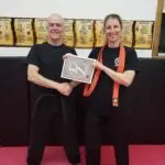 Two martial artists smile and shake hands on a pink mat, with a certificate being handed over as the woman wearing an orange belt stands beside the man.