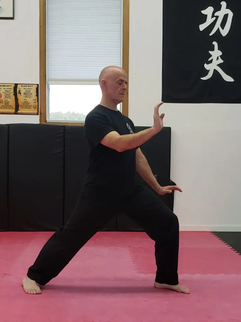 Bald man in black clothing performing a martial arts forward stance on a pink mat in a dojo.