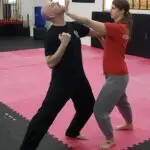 Two people spar in a dojo: a bald man in black with fists up while a woman in red presses his neck with her extended arm during a drill on pink mats.