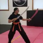 Person in black uniform and orange belt practicing martial arts, holding a silver sword in the left hand and a pink sword in the right, in a dojo.
