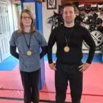 Two people wearing round medals stand side by side in a martial arts gym, barefoot on pink and blue foam mats.