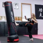 Young girl barefoot in a black uniform kicks a tall black and red punching bag in a martial arts gym as mats line the floor.