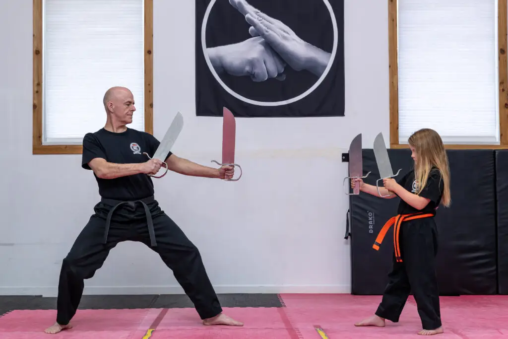Adult man and a young student practice sword fighting with large props on pink mats in a dojo, both wearing black uniforms and bare feet.