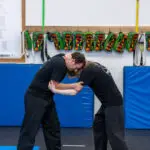 Two people grappling on padded gym mats, facing each other with heads close together during training.