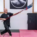 Bald man wearing a black martial arts gi practicing with a long spear in a dojo with padded mats.