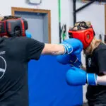 Two people sparring in a gym: one wears red headgear and blue gloves, throwing a punch toward the other.