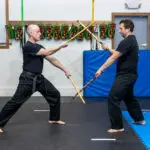 Two men in black martial arts uniforms spar with wooden sticks in a dojo, with colorful gear hanging on the wall behind them and blue mats on the floor.